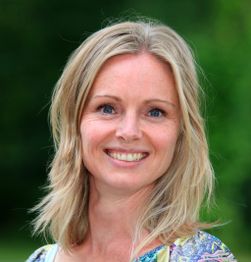 Een glimlachende vrouw met blond haar, buiten gefotografeerd met een groene achtergrond.
