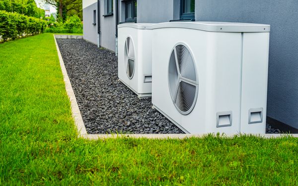 Eine Wärmpumpe auf einer Kiesfläche steht vor einem Mehrfamilienhaus.