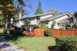 Two-story house with wooden fence, lush greenery, and a clear blue sky.