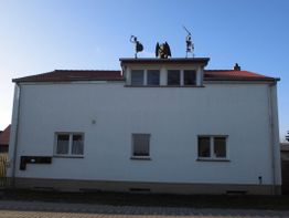 Zweistöckiges weißes Haus mit zwei Statuen und Antenne auf dem Dach unter blauem Himmel.