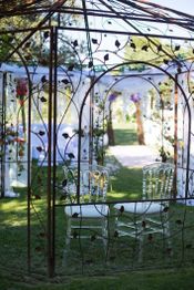 Deux chaises transparentes sous une arche décorée de fleurs dans un jardin ensoleillé.