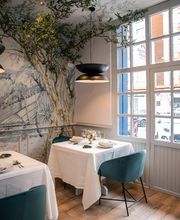 Restaurante elegante con mesas blancas, sillas verdes y mural de árbol en la pared. Ventanas grandes.