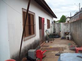 Quintal simples com paredes desgastadas, cadeiras, baldes e varal em frente a uma casa branca.