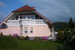 Rosa Haus mit Balkon, Garten und bewölktem Himmel im Hintergrund.