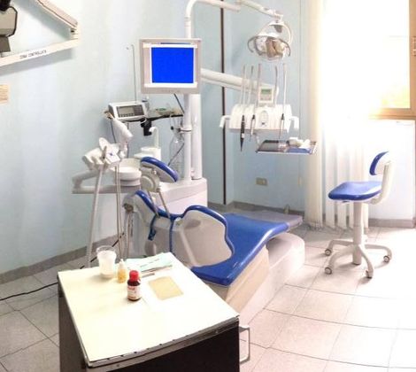 Studio dentistico con sedia blu, attrezzature odontoiatriche e monitor, illuminato dalla finestra.
