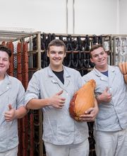 Drei Männer in Arbeitskleidung halten Wurst und Schinken, zeigen Daumen hoch, lächeln in einer Metzgerei.