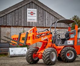 Orange Radlader vor einer Holzscheune mit mehreren Schildern, grauer Himmel im Hintergrund.