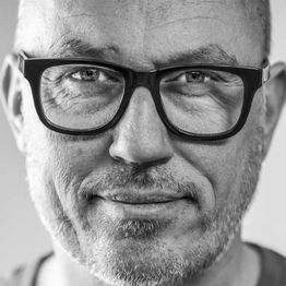 Graustufenfoto eines lächelnden Mannes mit Bart und großer Brille, direkter Blick in die Kamera.
