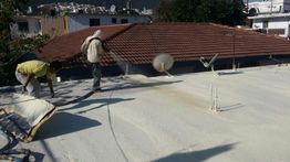 Trabajadores aplican recubrimiento en un techo plano, casas y árboles al fondo.