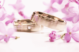 Zwei Eheringe, einer mit Diamant, umgeben von lila Blüten auf weißem Hintergrund.