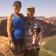 A couple and their dog smile on a scenic cliffside with a river and canyon in the background.