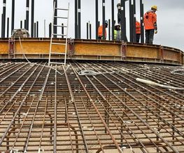 Bauarbeiter in orange Kleidung arbeiten auf einer Baustelle mit Stahlverstrebungen und Leitern.