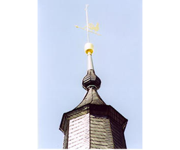 Kirchturmspitze mit Wetterhahn und goldener Kugel auf blauem Himmel.