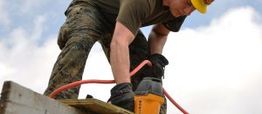 Worker in camo pants and helmet using a nail gun on a construction site.