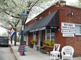 Brick building with canopy reads "Carpet & Blinds"; chairs and signs outside; tree in bloom nearby.
