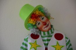 Ein Clown in buntem Kostüm und grünem Hut lächelt schief, vor weißem Hintergrund.