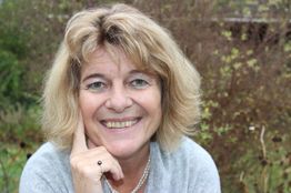 Eine lächelnde Frau mit blonden Haaren im Freien, trägt grauen Pullover und Perlenschmuck.
