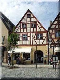 Fachwerkhaus an einem gepflasterten Platz, strahlend blauer Himmel, Passanten im Vordergrund.
