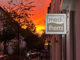 Ladenschild „medithera“ bei Sonnenuntergang in einer belebten Straße mit Bäumen und Passanten.