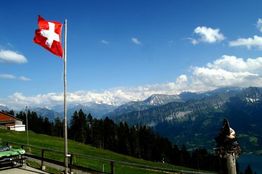Schweizer Flagge weht vor Alpenpanorama mit Berggipfeln und blauem Himmel im Hintergrund.