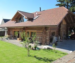 Blockhaus mit rotem Dach, Garten und kleinem Baum im Vordergrund. Sonniger Tag, blauer Himmel.