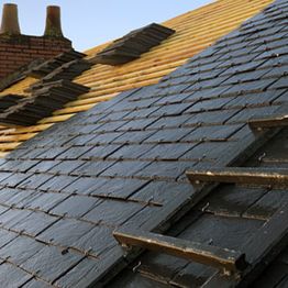 Toit en rénovation avec tuiles noires et jaunes, échelle posée dessus, cheminées en arrière-plan.