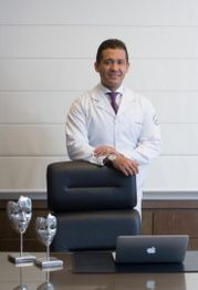 Homem de jaleco branco em pé atrás de uma cadeira, com um laptop e troféus na mesa.