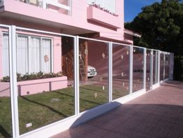 Casa rosa com cerca de vidro em frente, carro branco estacionado ao lado, céu azul ao fundo.