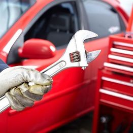 Gloved hand holding a wrench near a red car with a toolbox in the background.