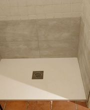 Ducha con plato blanco, azulejos grises, lavabo al lado y piso de cerámica marrón.