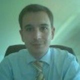 Ein Mann im Anzug sitzt und lächelt leicht, mit grünlichem Hintergrund.