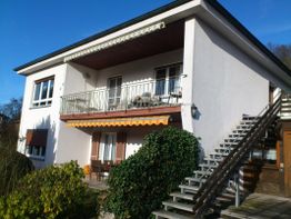 Zweistöckiges Haus mit Balkon und Außentreppe, umgeben von Grünpflanzen, unter blauem Himmel.
