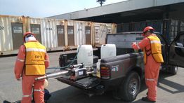 Trabajadores en overoles anaranjados preparan equipo de fumigación junto a un camión en un puerto.