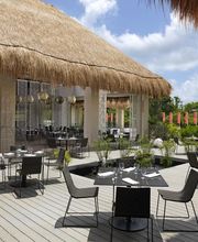 Restaurante al aire libre con techo de paja y mesas negras, rodeado de vegetación tropical.