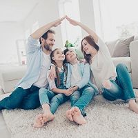 Family sitting on the floor at home, forming a roof shape with their arms, smiling together.