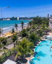 Praia paradisíaca com mar azul, piscina, coqueiros e prédio moderno ao fundo.