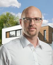 Ein Mann mit Brille vor einem modernen Haus, Bäume im Hintergrund, sonniger Himmel.