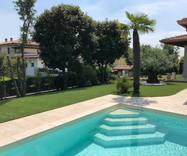 Garten mit Pool, Palmen und Rasenfläche, umgeben von Bäumen und modernem Haus bei klarem Himmel.