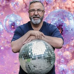 Mann mit Bart hält eine große Discokugel, im Hintergrund schwebende Discokugeln vor rosa Hintergrund.