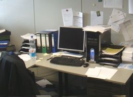 Unordentlicher Schreibtisch mit Computer, Ordnern, Flaschen und Papierstapeln.