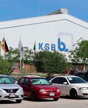 Fábrica con el logo KSB y banderas, estacionamiento con varios coches en frente.