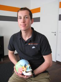 Mann in schwarzem Polohemd hält ein buntes Schädelmodell in einem Raum mit weißen Wänden.