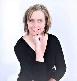 Eine lächelnde Frau in schwarzer Kleidung vor einem weißen Hintergrund, leicht seitlich sitzend.