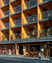 Mehrstöckiges Gebäude mit Balkonen, darunter ein Café mit Sitzbereich auf der Terrasse bei sonnigem Wetter.