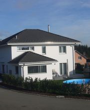 Zweistöckiges weißes Haus mit blauem Pool im Garten, umgeben von Hecke. Klarer Himmel im Hintergrund.