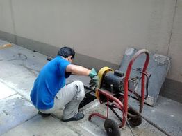 Homem trabalha com máquina de desentupimento em calçada, usando luvas verdes.