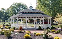 White gazebo in a park, surrounded by flowers and trees, under a clear sky.