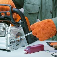 Person in orange gloves repairing a chainsaw with a tool.
