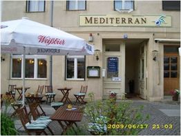 Außenansicht eines mediterranen Cafés mit Sitzbereich und Sonnenschirm auf einem Platz.