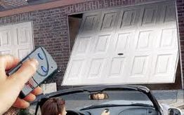 A person uses a remote to open a white garage door while sitting in a car.